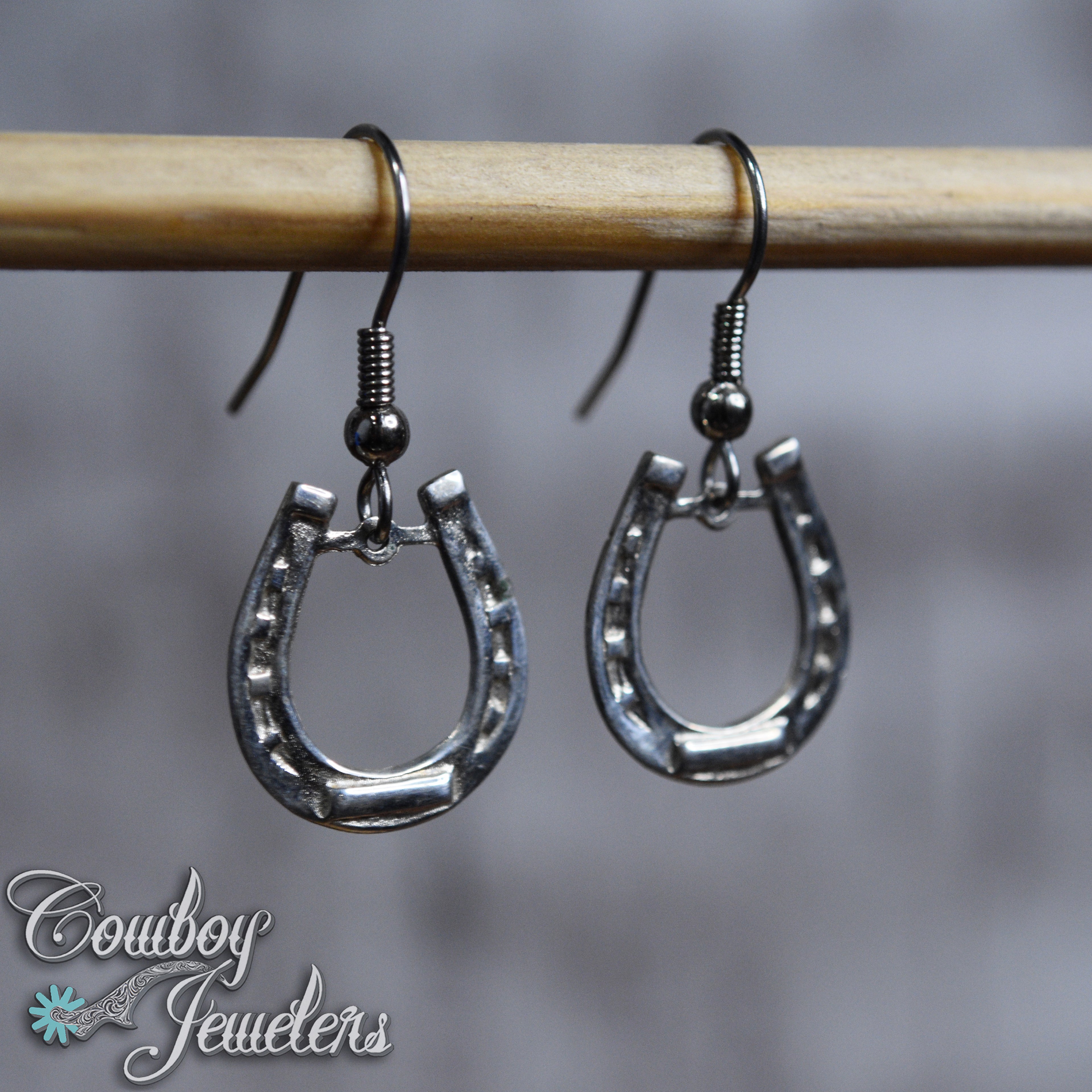Silver horseshoe earrings hanging on a rod with a blurred background and 'Cowboy Jewelers' branding.