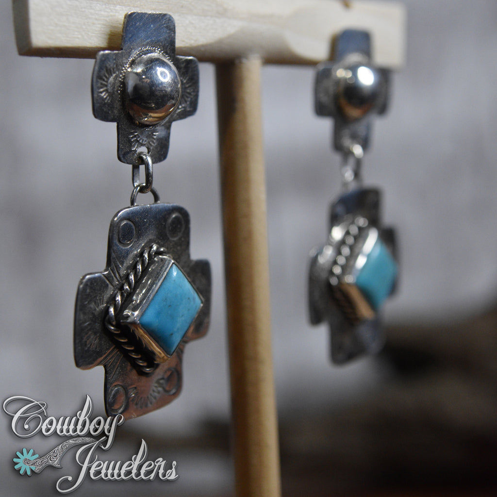 Pair of silver and turquoise earrings on a wooden stand with 'Cowboy Jewelers' branding.