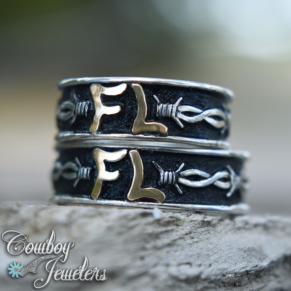 Stack of silver rings with barbwire designs on a stone surface, featuring the brand 'Cowboy Jewelers'.