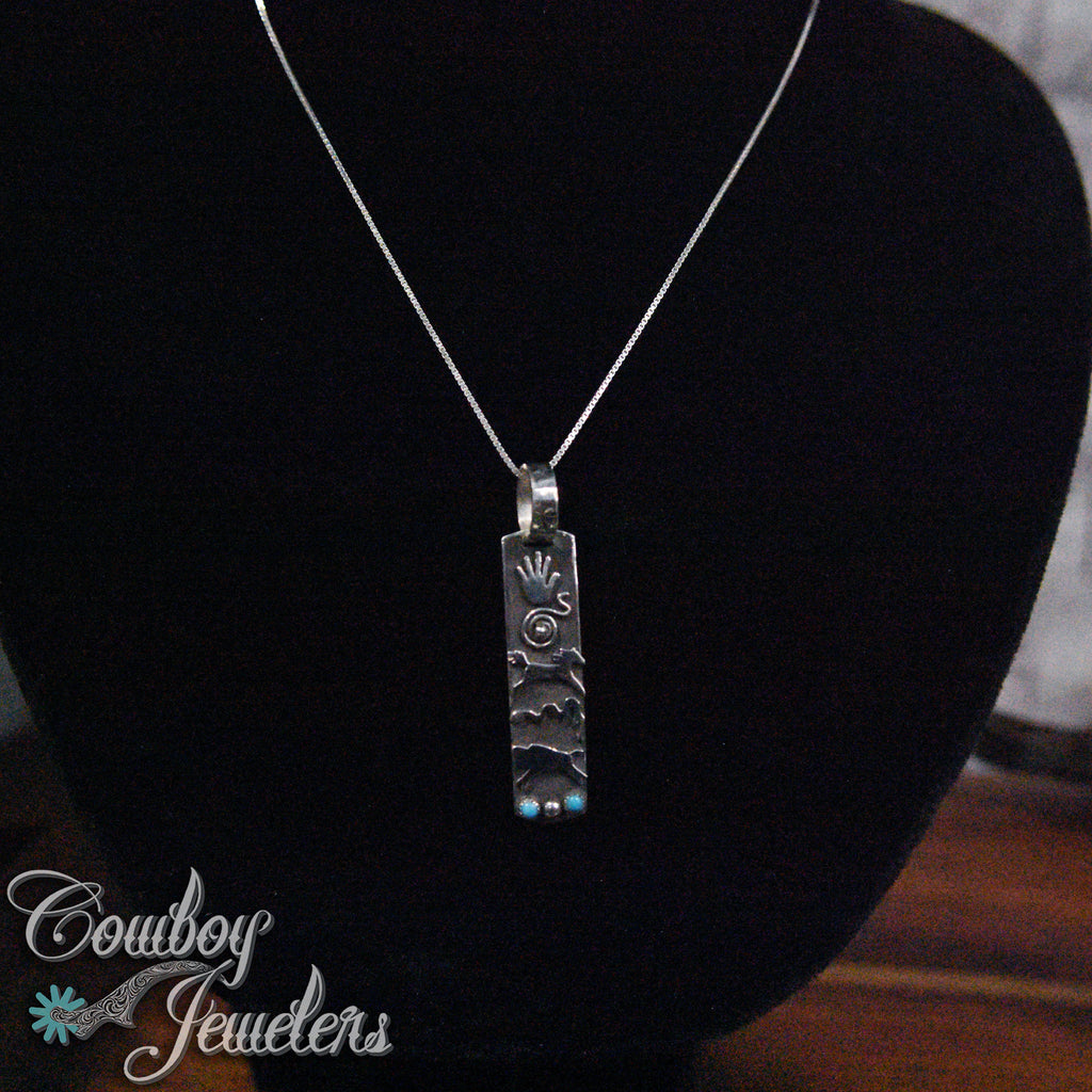 Silver pendant necklace with native American emblems on a black stand with 'Cowboy Jewelers' branding.