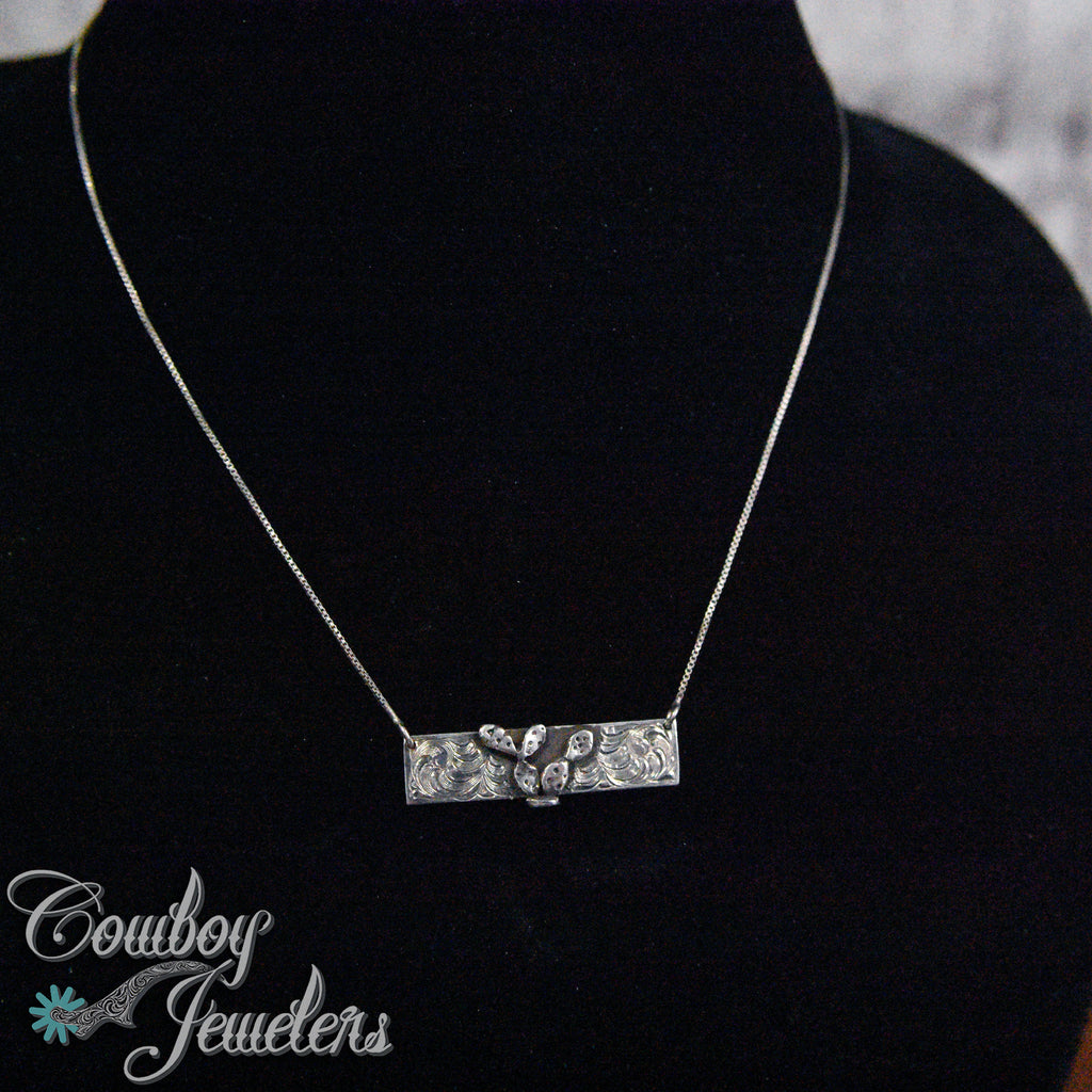 Silver necklace with a cactus pendant on a black stand, branded 'Cowboy Jewelers'.