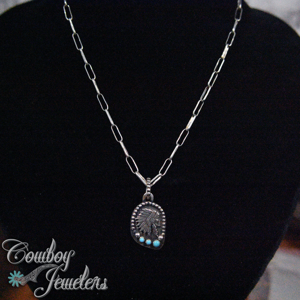 Silver Indian head  necklace with a pendant on a black stand, branded 'Cowboy Jewelers'.