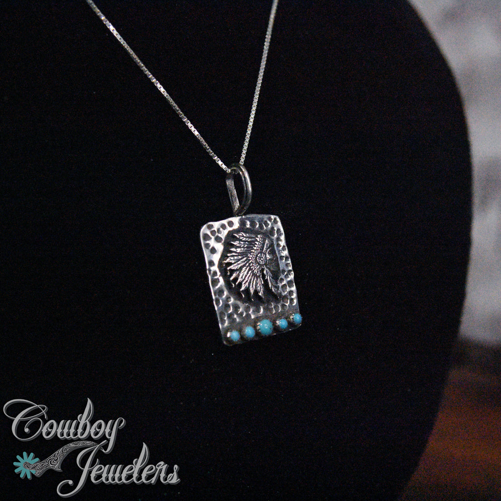 Silver Indian head pendant and  necklace with turquoise stones on a black stand, branded 'Cowboy Jewelers'.