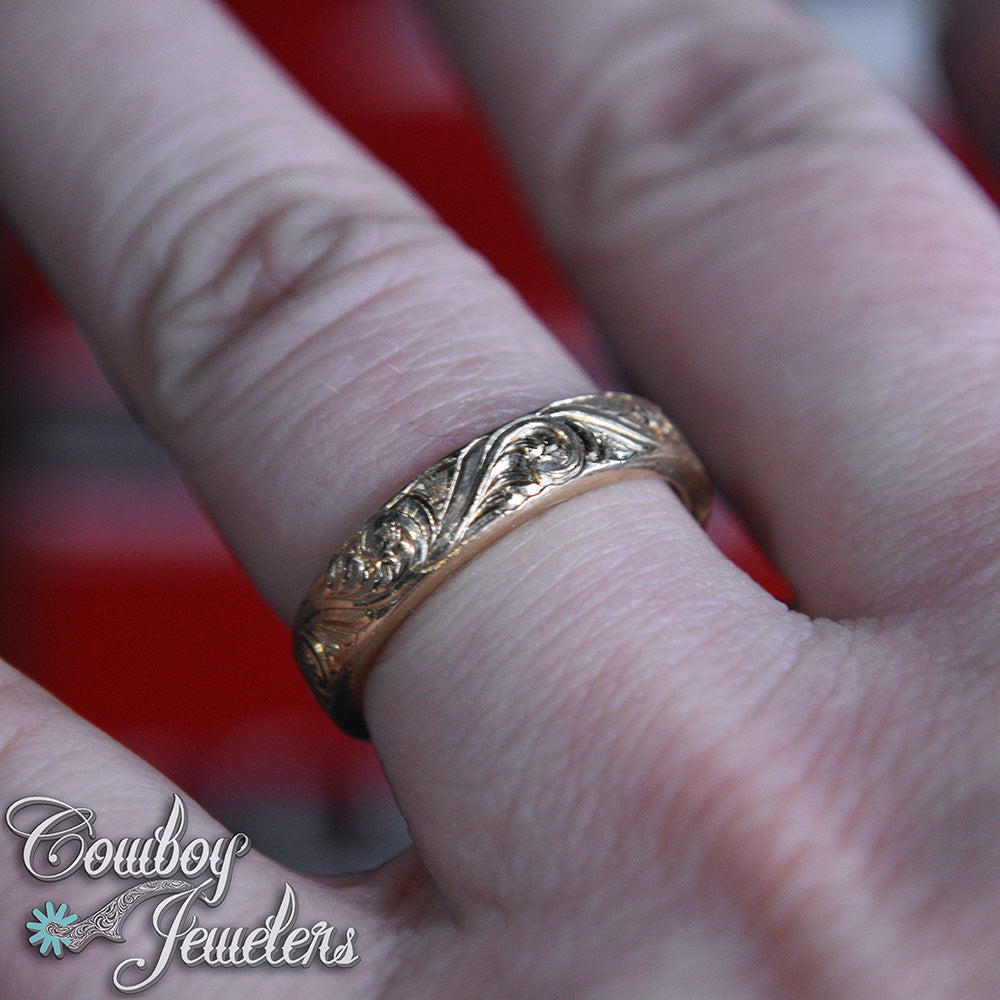 Gold engraved  ring on a finger with 'Cowboy Jewelers' branding.