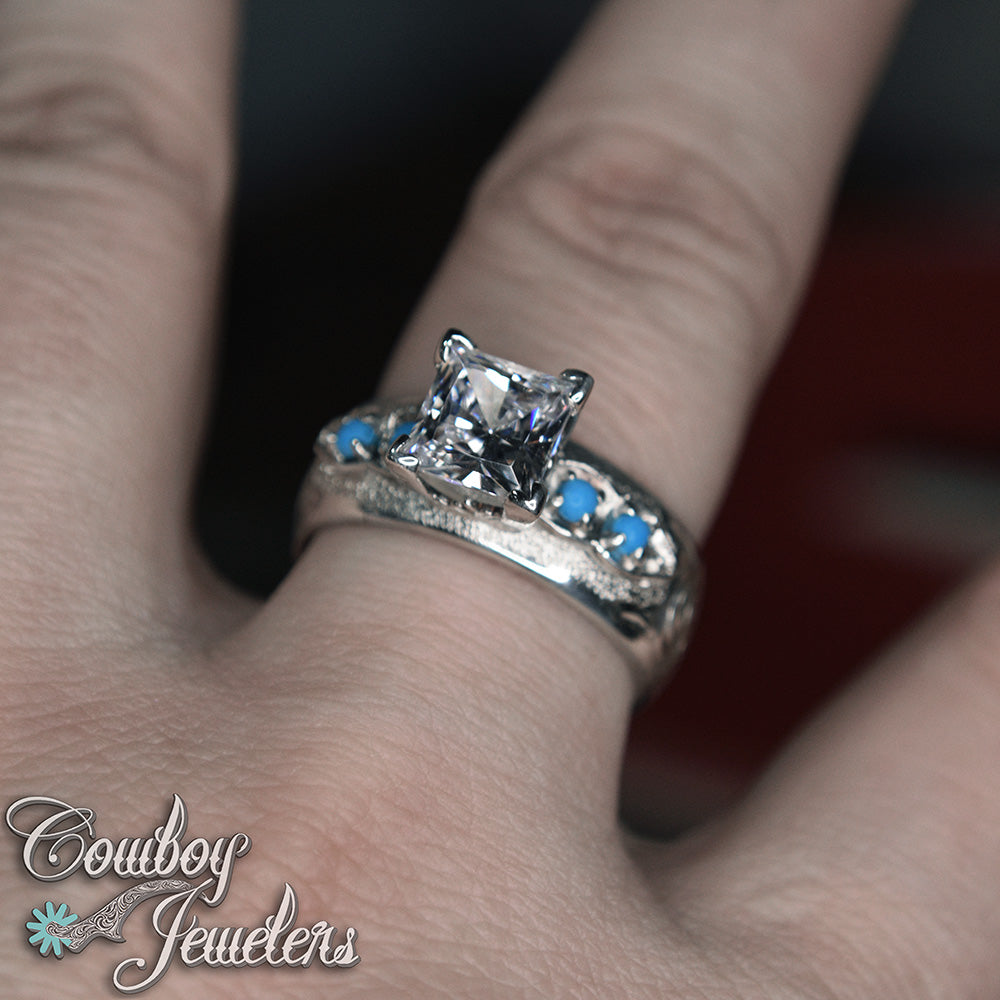 Silver ring with a large central gemstone and smaller stones on a finger, branded 'Cowboy Jewelers'.