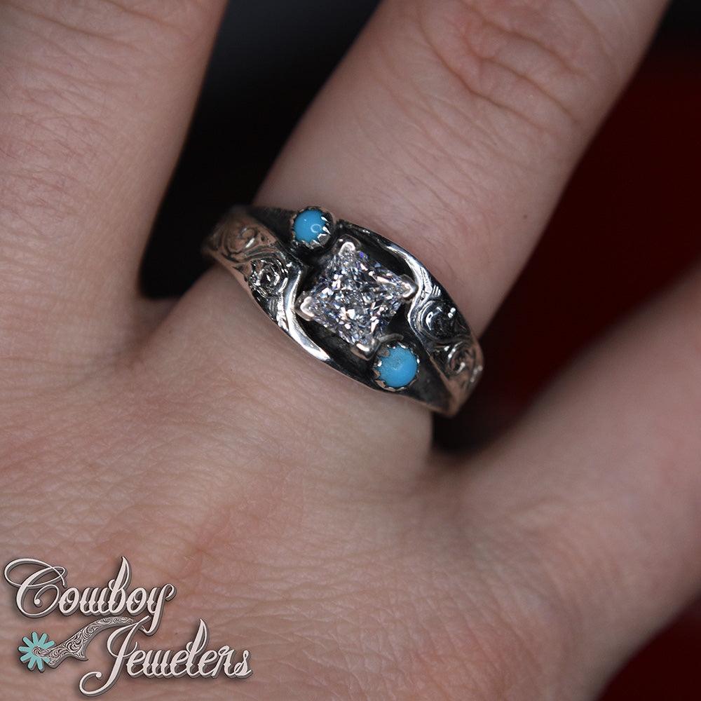 Silver ring with a diamond and turquoise stones on a finger, branded 'Cowboy Jewelers'.