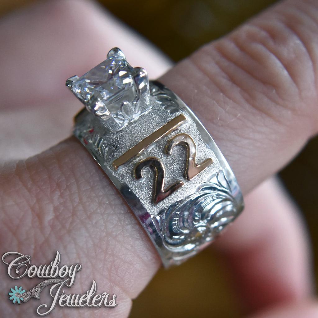 Silver ring with custom brand and a  diamond on a finger, branded 'Cowboy Jewelers'.