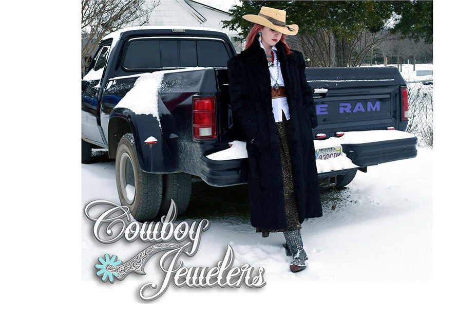 Person in cowboy hat and coat standing next to a black Ram truck in a snowy setting, with 'Cowboy Jewelers' branding.