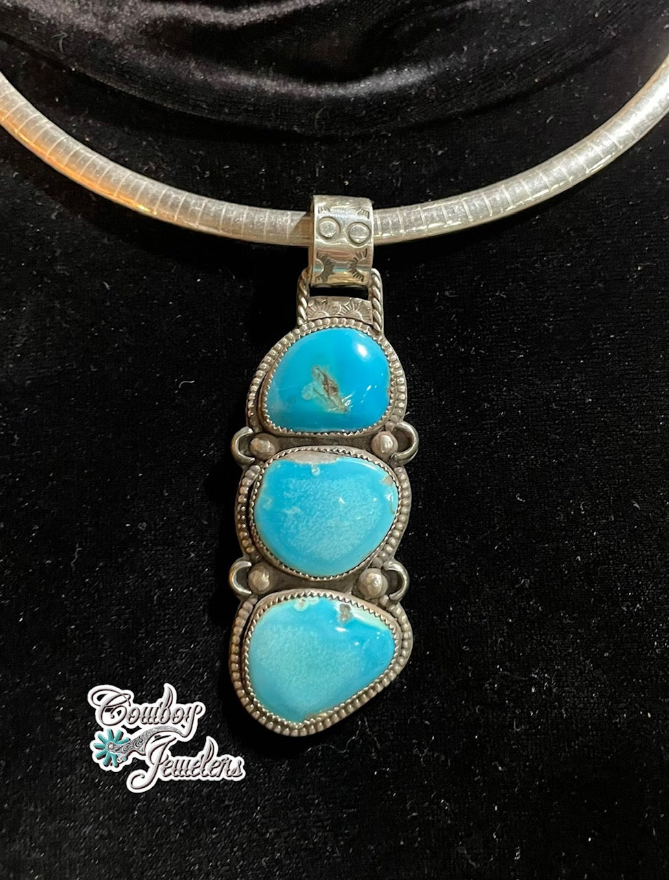 A silver-toned herringbone necklace with a vertical three-stone Kingman turquoise pendant, displayed on a black velvet surface. A 'Cowboy Jewelers' watermark is visible in the lower-left corner.