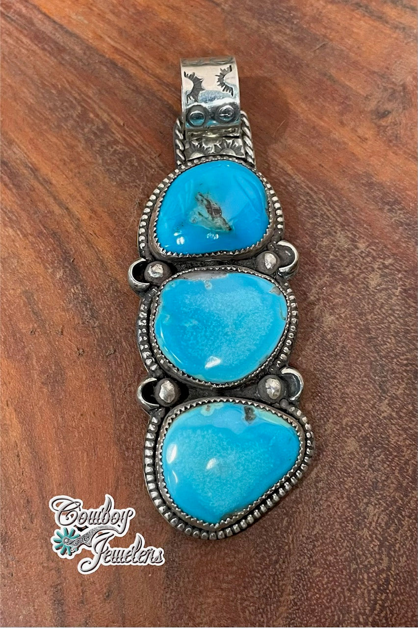 A Southwestern-style sterling silver pendant featuring three stacked oval kingman turquoise cabochons, displayed on a dark brown wooden surface. The silver bezel settings are detailed with small silver dots and a decorative bail at the top.