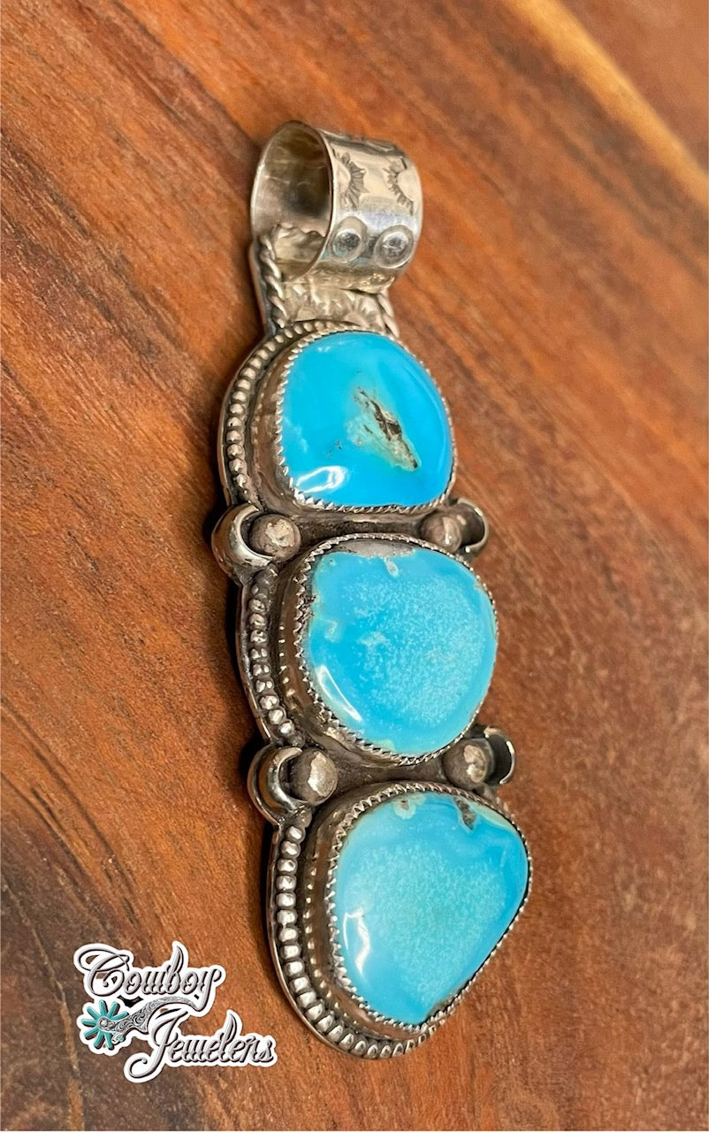 A side view of a sterling silver pendant featuring three stacked oval kingman turquoise stones, displayed on a warm brown wooden surface. The brand logo 'Cowboy Jewellers' is visible in the bottom-left corner.