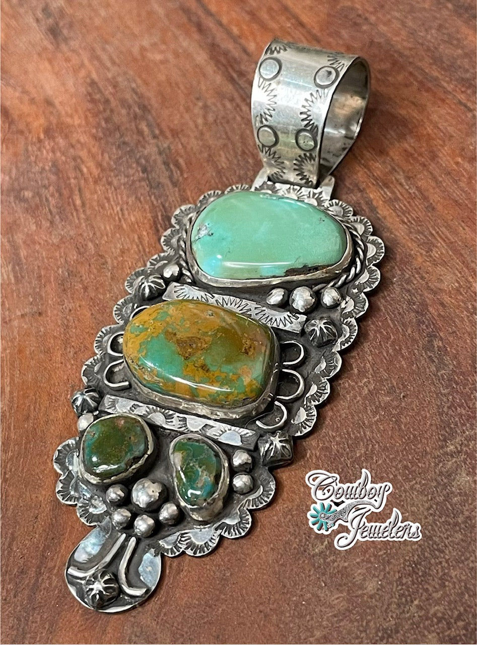 A side view of a sterling silver pendant featuring 2 large royston turquoise stones in a native american style setting, displayed on a dark wood background. A 'Cowboy Jewellers' logo is visible in the bottom right corner.