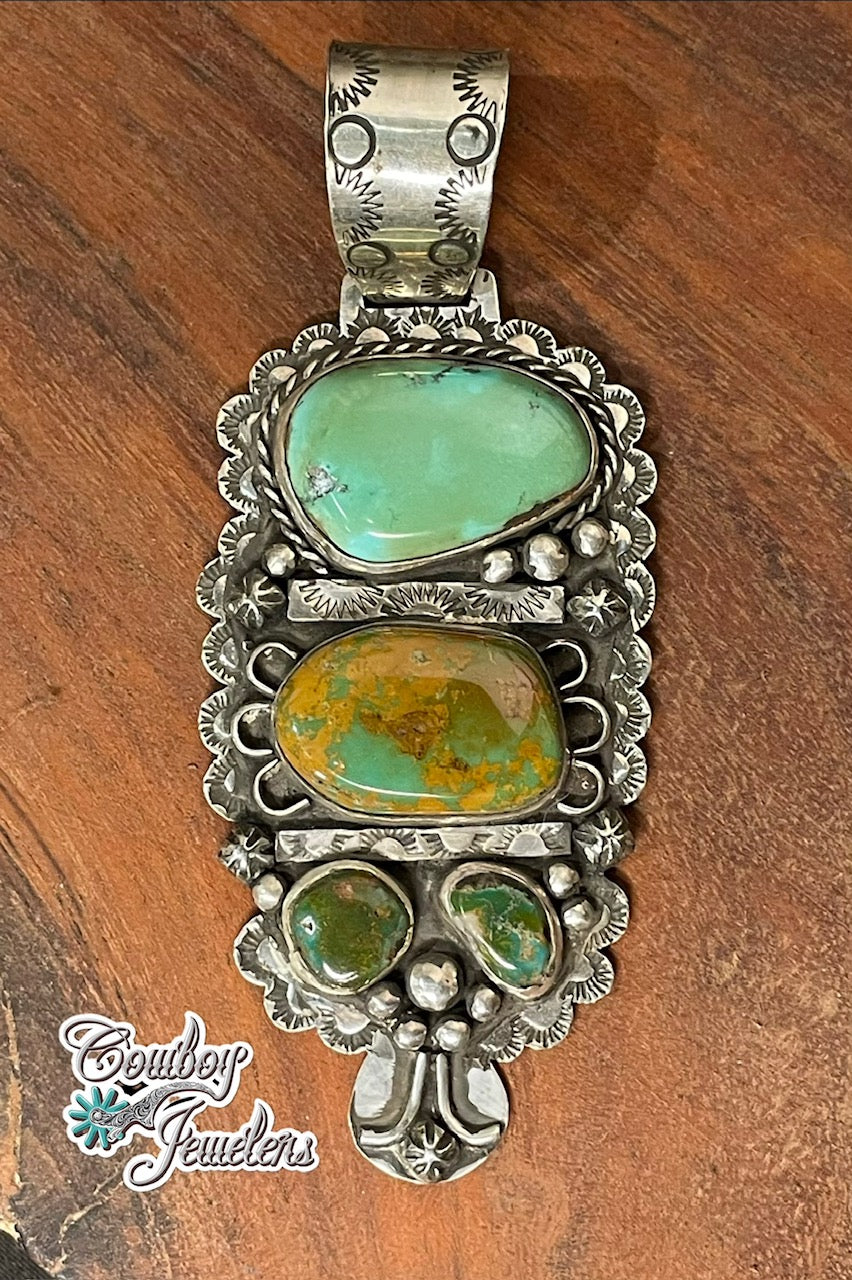 A detailed silver-toned Southwestern-style pendant featuring royston turquoise stones, displayed on a wooden surface with a 'Cowboy Jewellers' watermark in the lower-left corner.